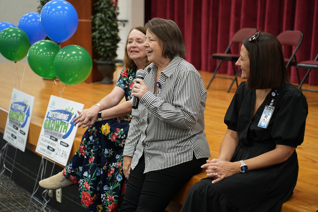 Three women seated on a stage with balloons behind them; one holding a microphone, the others smiling