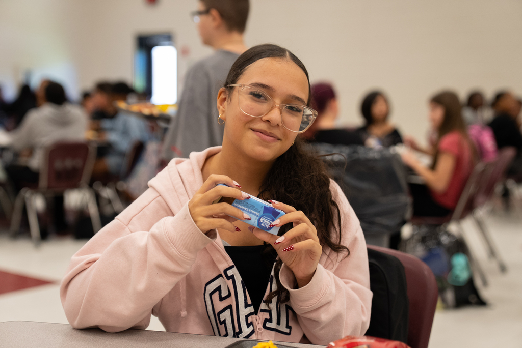 A girl with glasses holds a blue object in her hands while sitting in a cafeteria.
