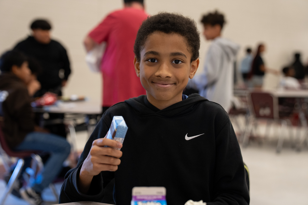 A child in a black hoodie smiles while holding a blue box. Behind them are other people seated at tables.