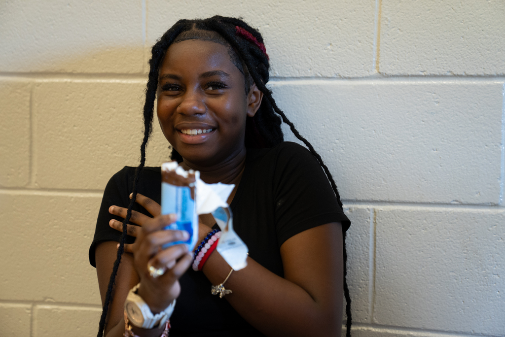 A girl with long braids holds a chocolate bar and smiles, seated against a white brick wall.