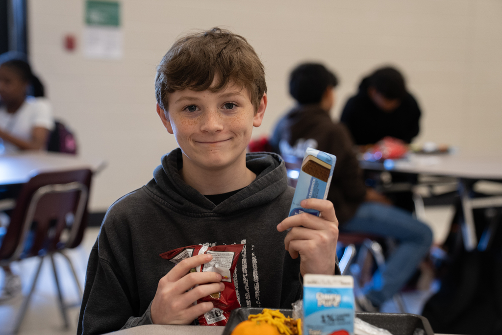A boy in a black hoodie holds a chocolate bar, chips, and a carton of milk.