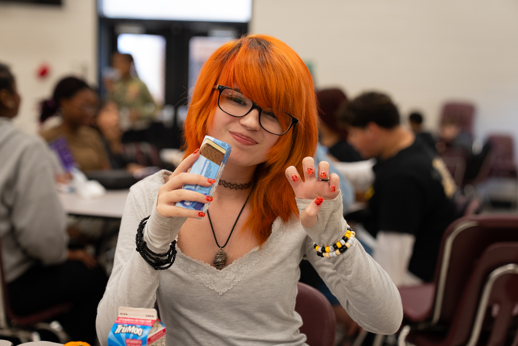 A student with red hair, glasses, and painted fingernails holds a blue box with a food item.