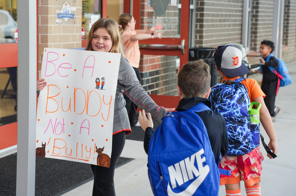 Girl holds sign with text "Be a Buddy, Not a Bully". Boy hugs girl. Children walk with backpacks.