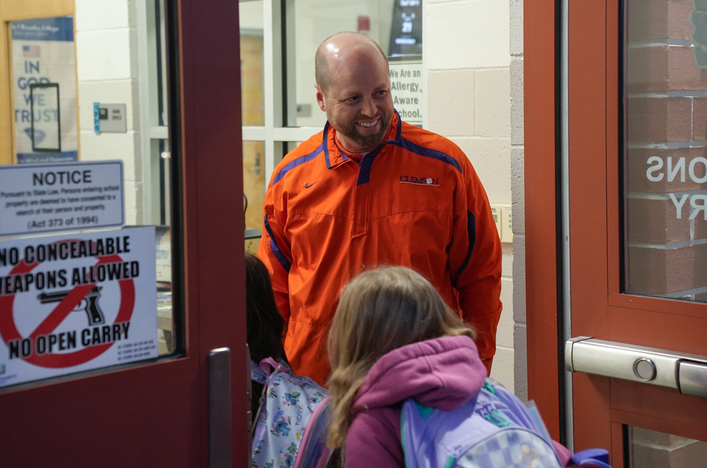 Man in orange coat stands at door with two students, one with backpack, and sign prohibiting concealed weapons.