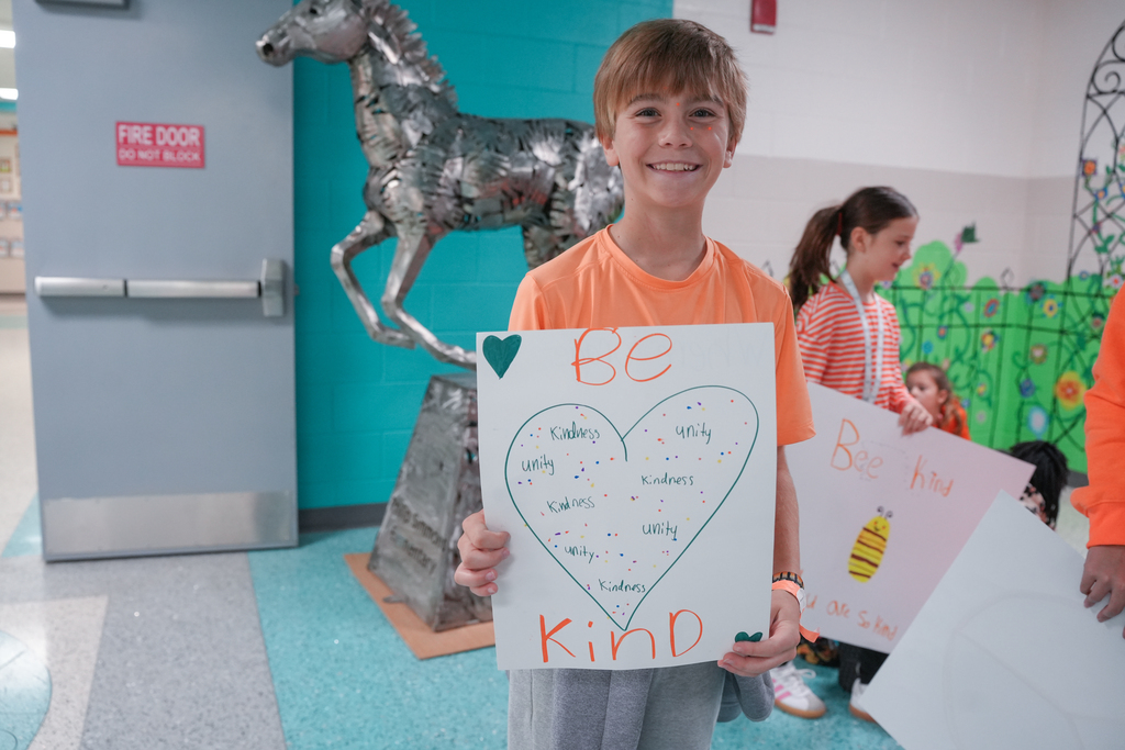 Boy in orange shirt holding sign that says "Be Kind" with heart on it.