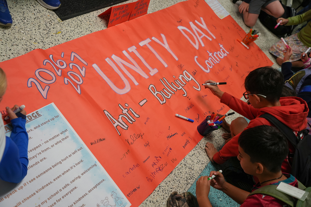 Several children sit cross-legged, writing on a large orange banner with white marker. The banner says "2017 Unity Day" and "Anti-Bullying Contract"