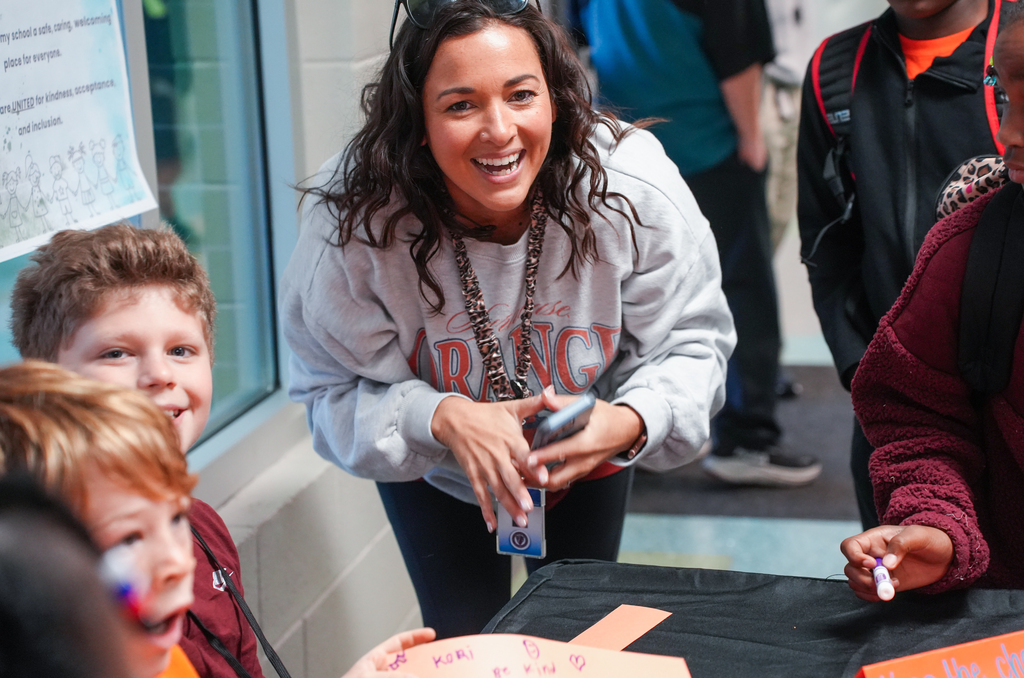 Woman in gray sweatshirt with text standing by table, smiling at four children; table has paper cutouts and writing utensils.