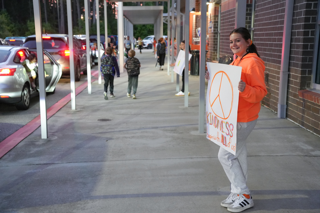 Woman in orange jacket holding a peace sign, standing next to school building as children and cars line street.