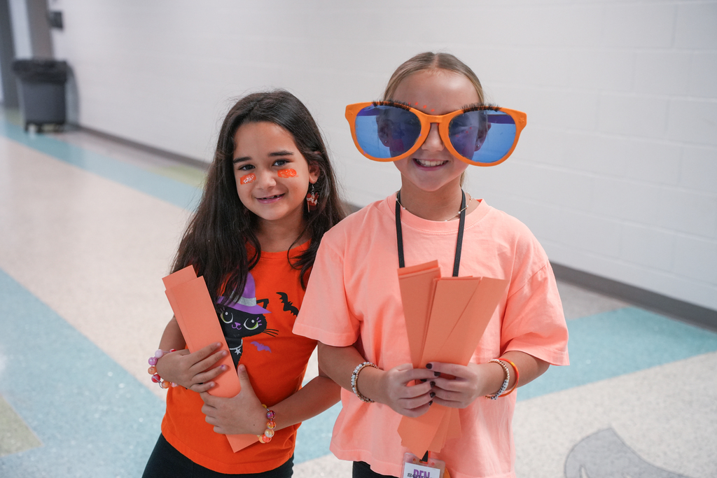 Two young girls in orange shirts stand in a hallway, one with face paint and both holding orange papers.