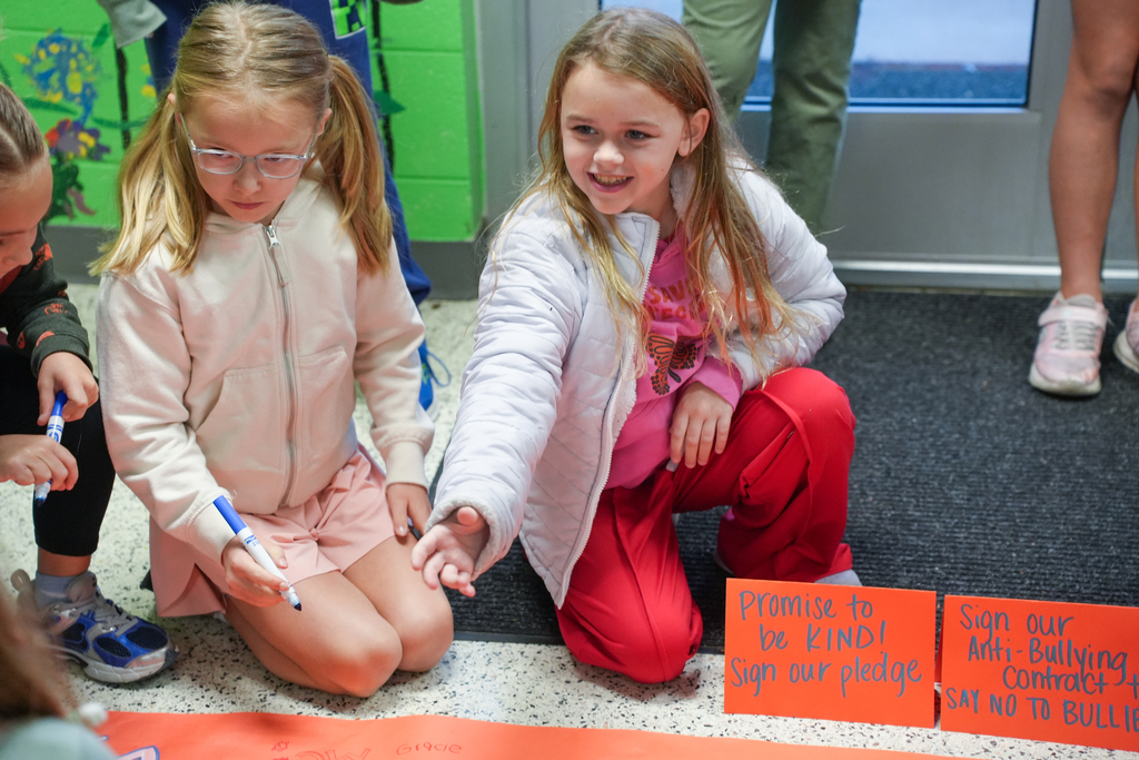 Three young girls sit on a floor, each holding a marker, writing on orange banners. One banner reads "Promise to be kind!" Another says, "Sign our pledge!"