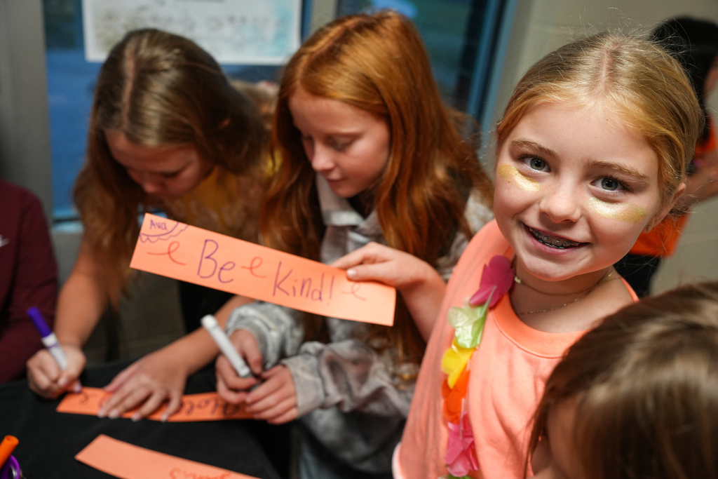 Four young girls sit around a table with markers and paper, two hold signs that say "Be Kind."