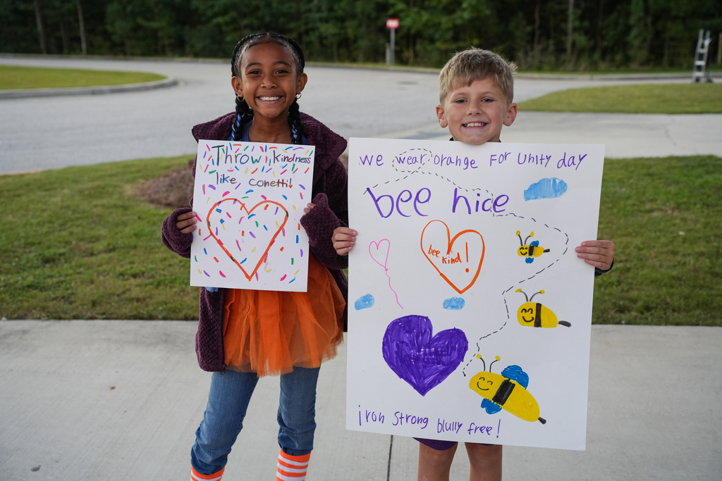 Two children stand on the curb holding signs with heart-shaped drawings, one with a bee and a path.