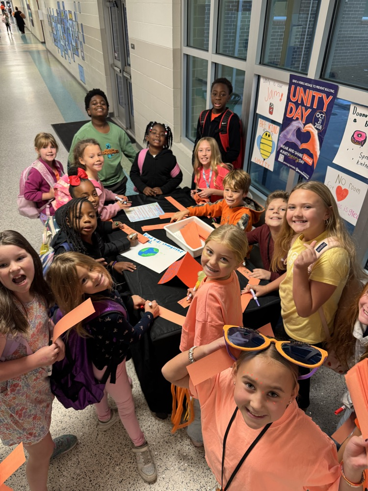 Group of children stand in a hallway around a table holding pieces of paper, some wear sunglasses.
