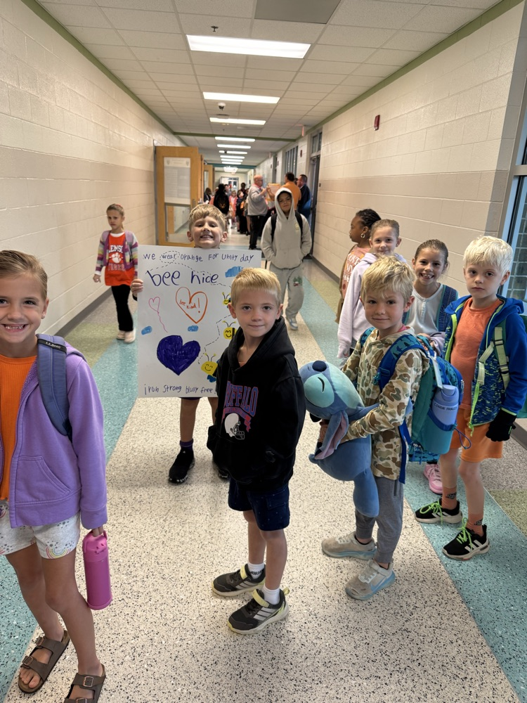Children wearing backpacks stand in a hallway holding a sign that says "bee hive"