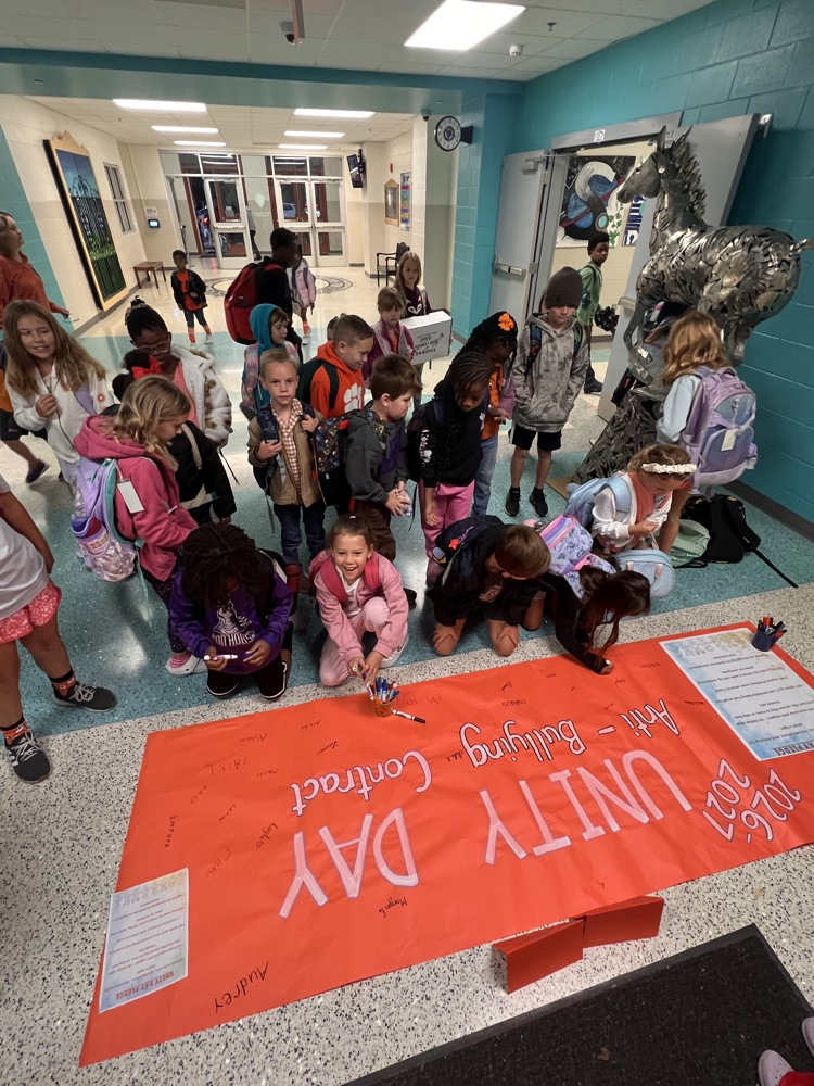 Children and adults in a school hallway. Children kneeling on the floor around a large banner.