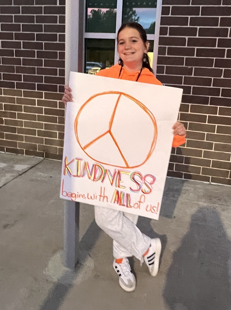 A girl holds a sign with a peace symbol and the words "Kindness begins with all of us!"