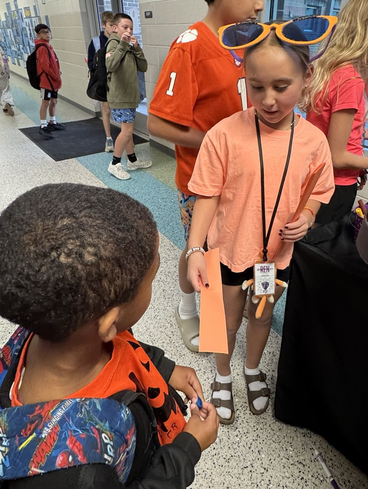 A group of children in a school hallway, one holding a pencil and a piece of paper.