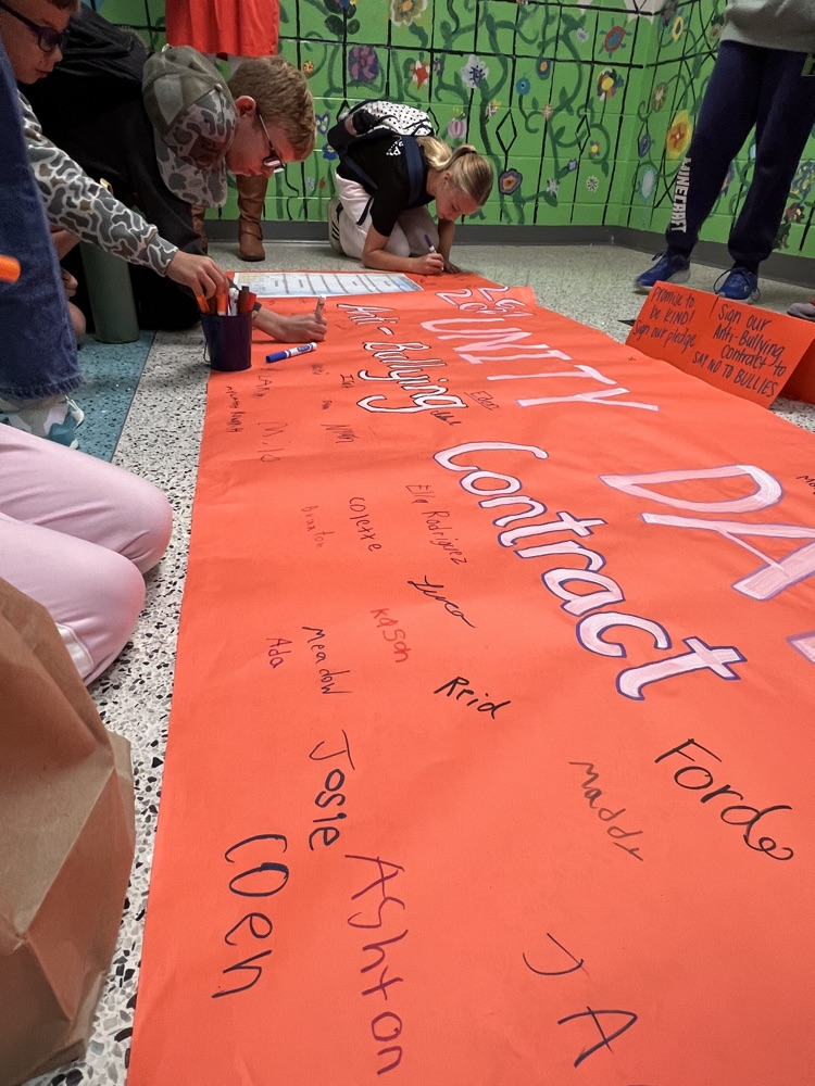 Five students wearing backpacks kneel to write on an orange banner on the floor, with many names already present.