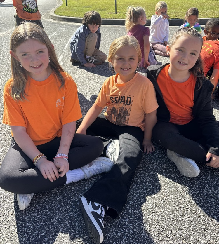 Children sitting on ground, three wearing orange shirts, one with black pants, another with white shoes.