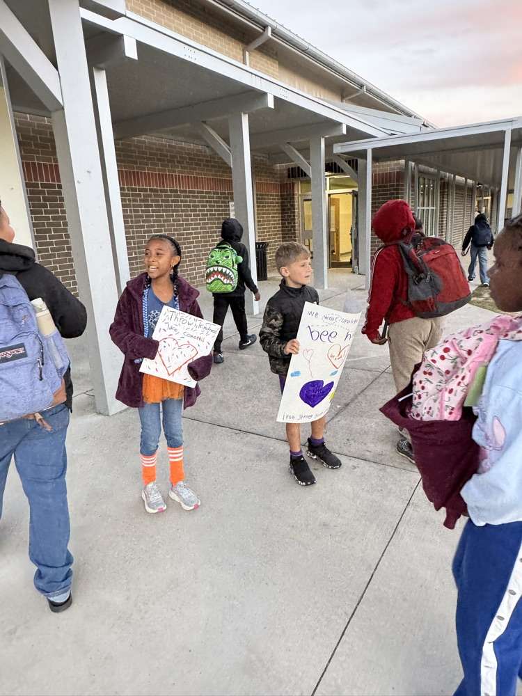 Several children stand under covered school walkway, holding signs and backpacks, with others walking in the background.