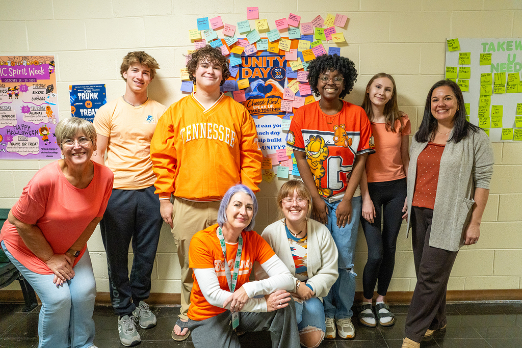 Nine people stand and crouch in front of a wall with several colorful notes pinned to it.