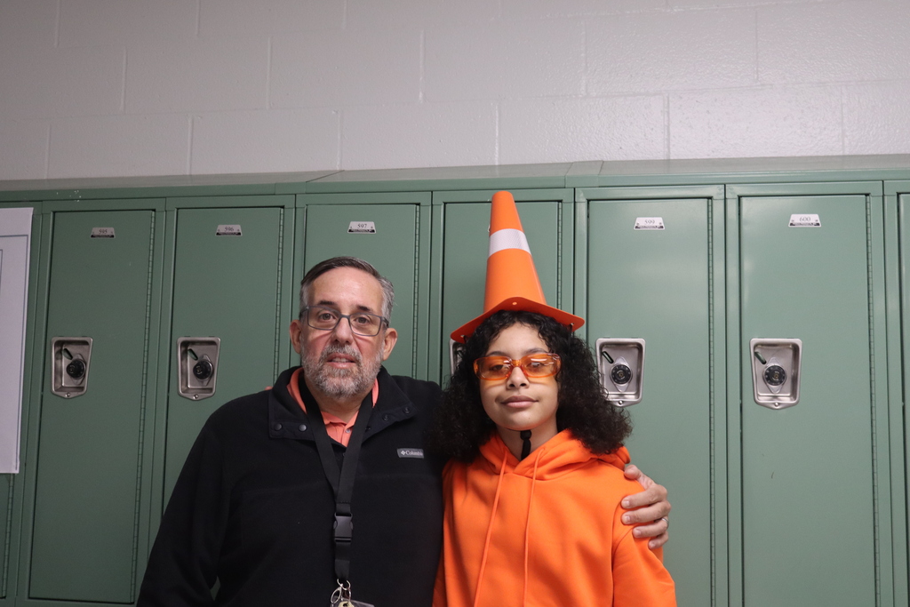 Man and girl stand in front of lockers; man wears black jacket and orange cone on head.