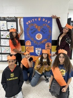 Group of children in orange crowns posing with a blue banner in a classroom.