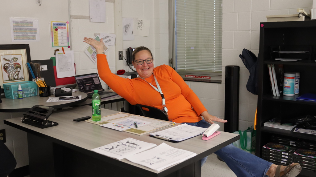 A smiling woman in glasses sits at a desk in an office, gesturing with her arm.