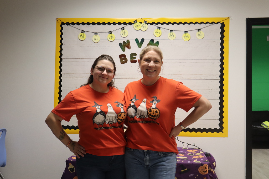 Two women in orange shirts stand next to each other in front of a bulletin board.