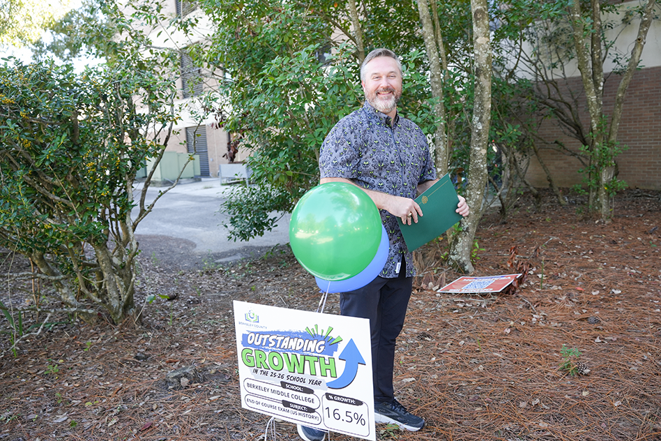 Person in black pants and sneakers holds green balloon and green folder in outdoor area. Sign with green arrow and text.