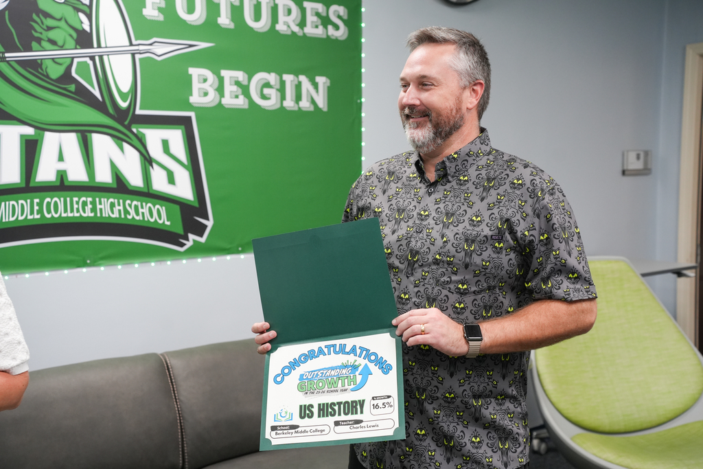 A smiling man holding a certificate, wearing a watch, stands in a room with a green banner.
