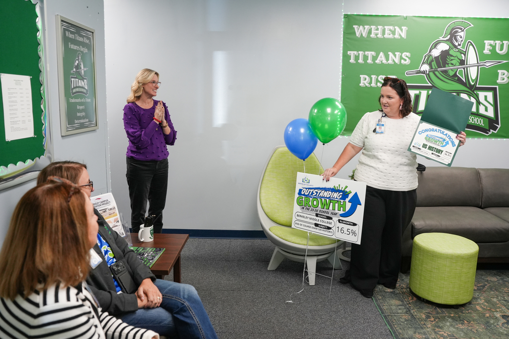 People seated around a table listen to a woman holding a folder. Two other women stand, one holding a green balloon and a sign.