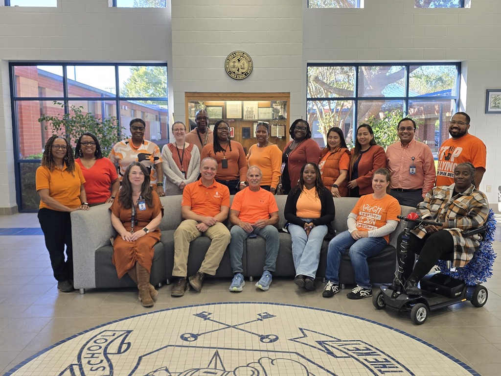 A group of adults and children in orange shirts pose for a photo in a hallway.