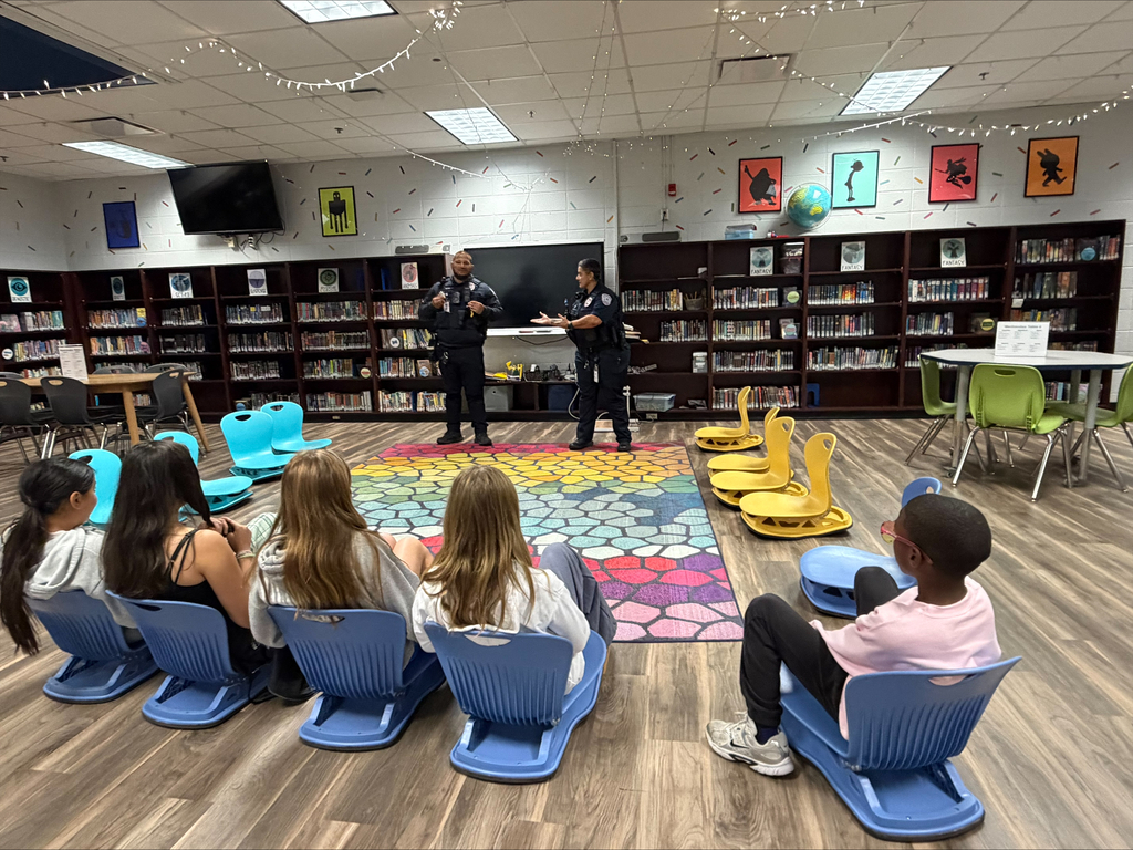 Two police officers stand by a colorful rug in a library. Several students sit around the rug on chairs.