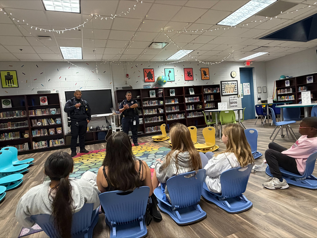 Police officer stands in front of students sitting in chairs with colorful mats on floor.