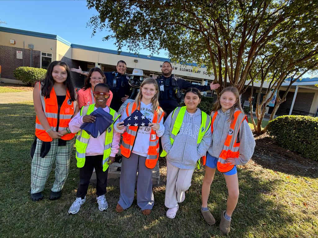 Several students in vests stand on a lawn. A girl holds an American flag. A building and trees are in the background.