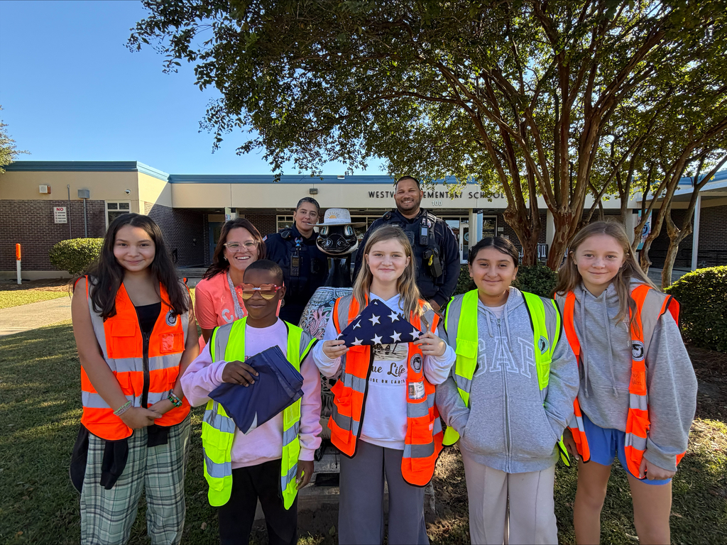 Group of children and adults in safety vests standing in front of a building with trees.
