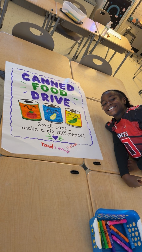 Student smiling beside a banner for canned food drive. Banner displays canned food with text "Canned Food Drive, Small Cans... Make a Big Difference!" Basket of markers nearby.