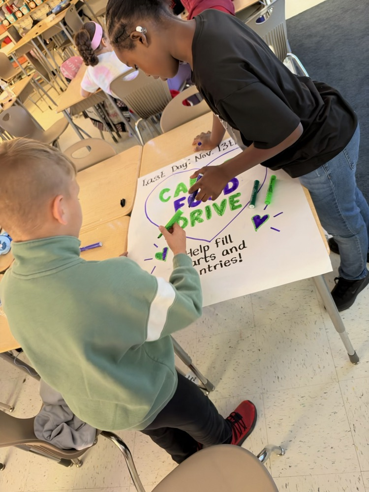 Two students work on a white poster with blue and green markers. The poster says, "CAN FOOD DRIVE" and "Help Fill Carts and Entries".