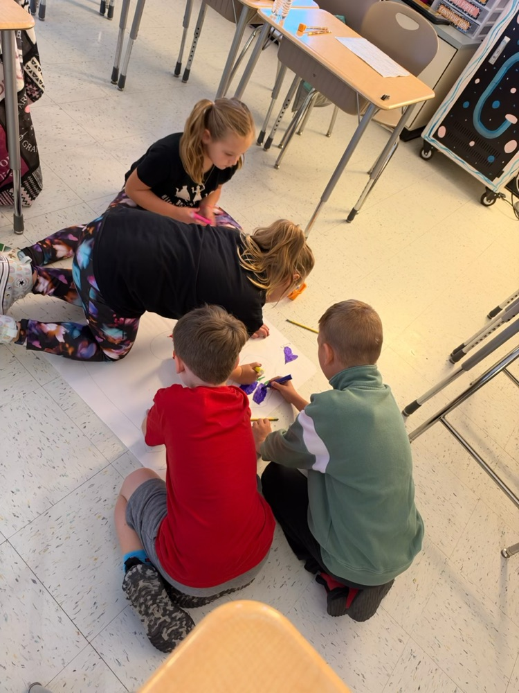 Students sit on floor around paper, drawing with markers; teacher assists. Behind them, tables and chairs.