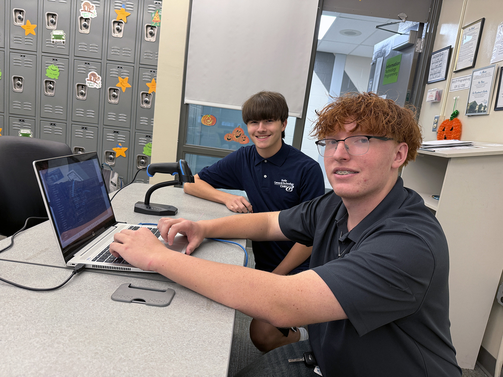Two IT Programming students sit at a classroom table working on a laptop. In the foreground, Chase, a student with red hair and glasses, types on the keyboard while looking toward the camera. Next to him sits Michael, with straight brown hair, smiling as he faces forward. A barcode scanner is positioned beside the laptop. Behind them are gray lockers decorated with fall-themed cutouts.
