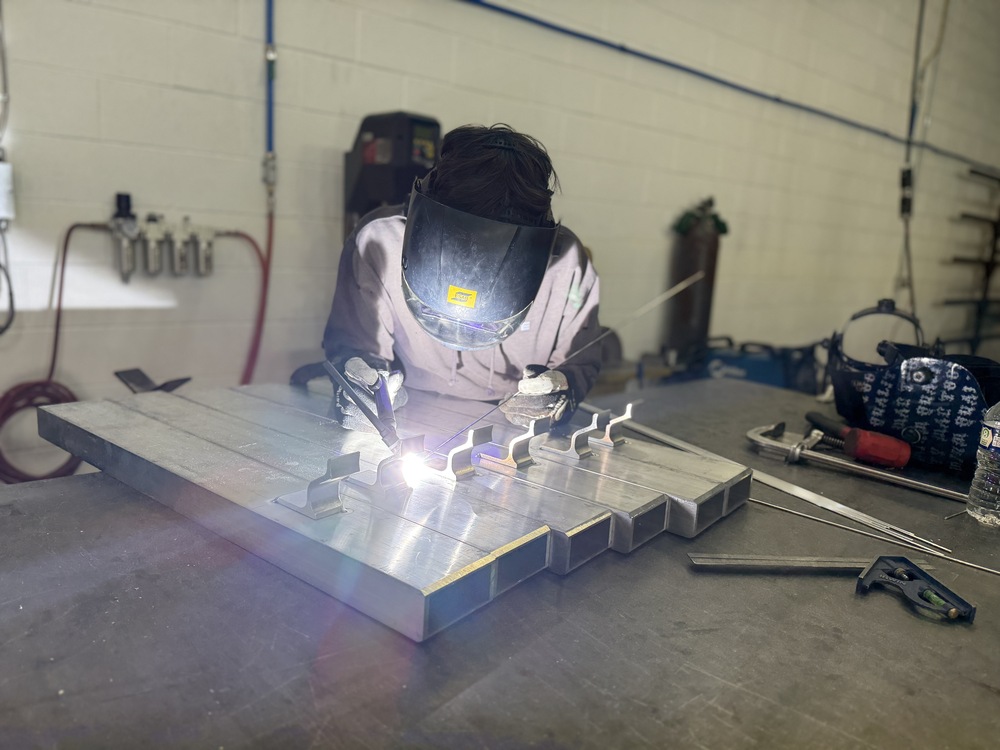 A student wearing a welding helmet welds metal pieces on a worktable in a career and technical education lab.