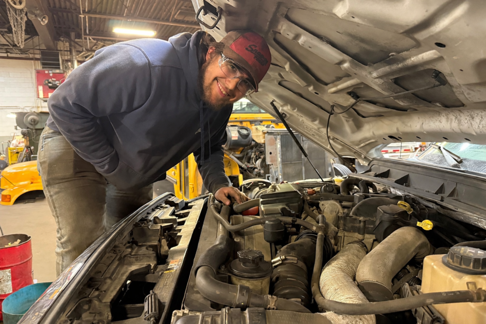 Student wearing safety glasses works under the hood of a vehicle, inspecting engine components in an auto shop.