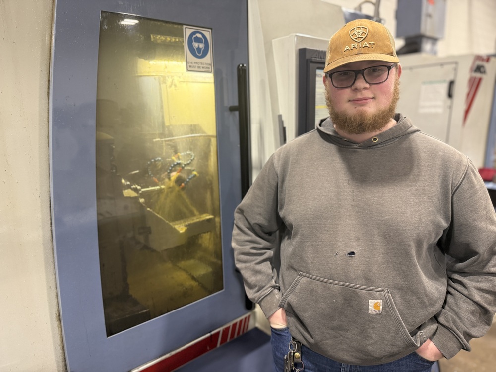 A student wearing an Ariat hat and gray hoodie stands beside a large piece of shop equipment in a career and technical education lab.