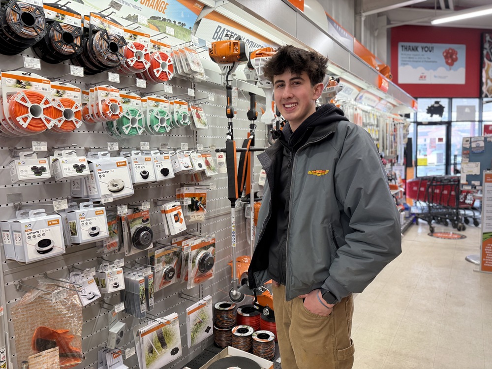 Student stands in a hardware store aisle, smiling beside a display of trimmer parts and lawn equipment accessories.