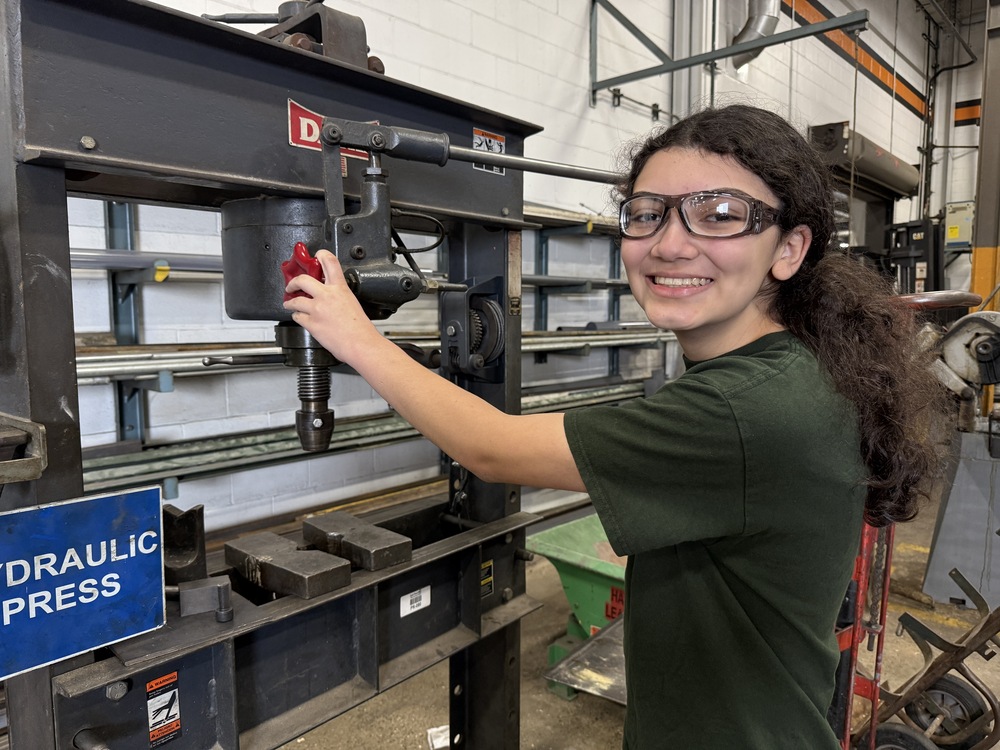 Mechatronics Engineering Technology student Pam Guzman smiles while wearing safety glasses and working with a hydraulic press during her co-op experience at East Penn Manufacturing.