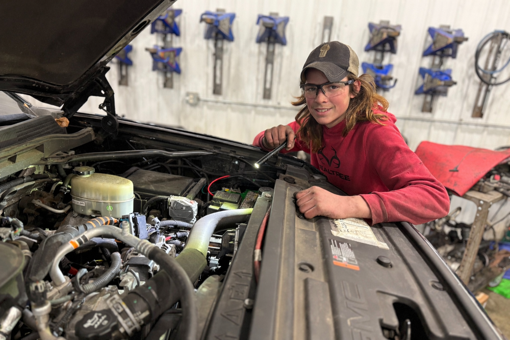 Student wearing safety glasses inspects a truck engine with a flashlight in an automotive lab, leaning over the open hood.