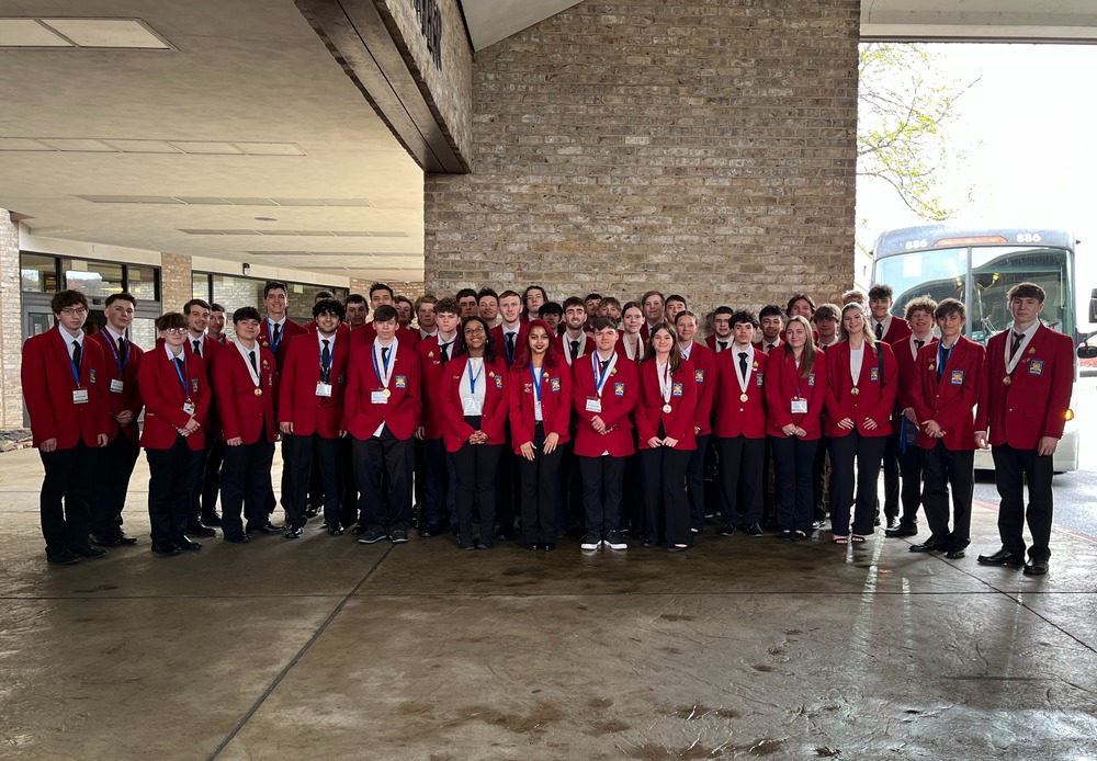 A group of students dressed in red suit jackets, black pants, and white shirts, standing in front of a brink wall