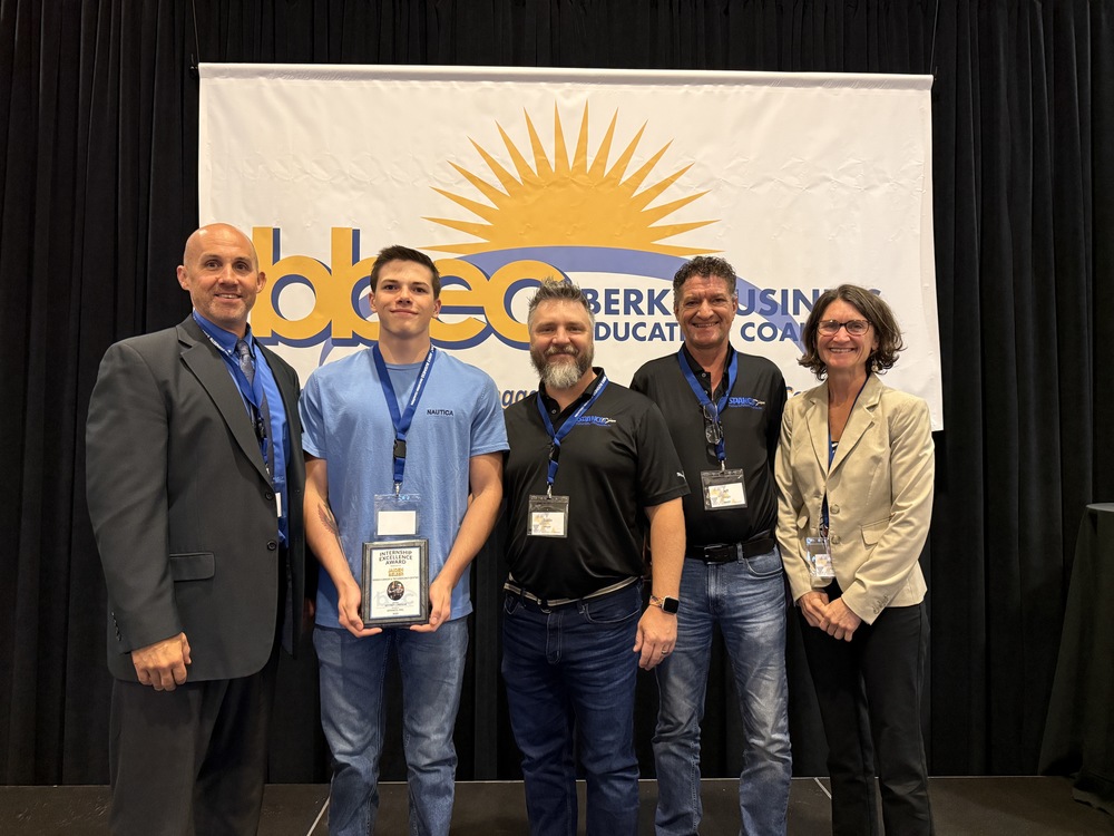Five individuals stand together in front of a banner that reads “Berks Business Education Coalition.” In the center, Welding Technology student Jaiden Belser holds a framed award. He is joined by representatives from BCTC and Spanco PTP, all smiling and dressed in professional or business-casual attire. The group is positioned on a stage with a black curtain backdrop.