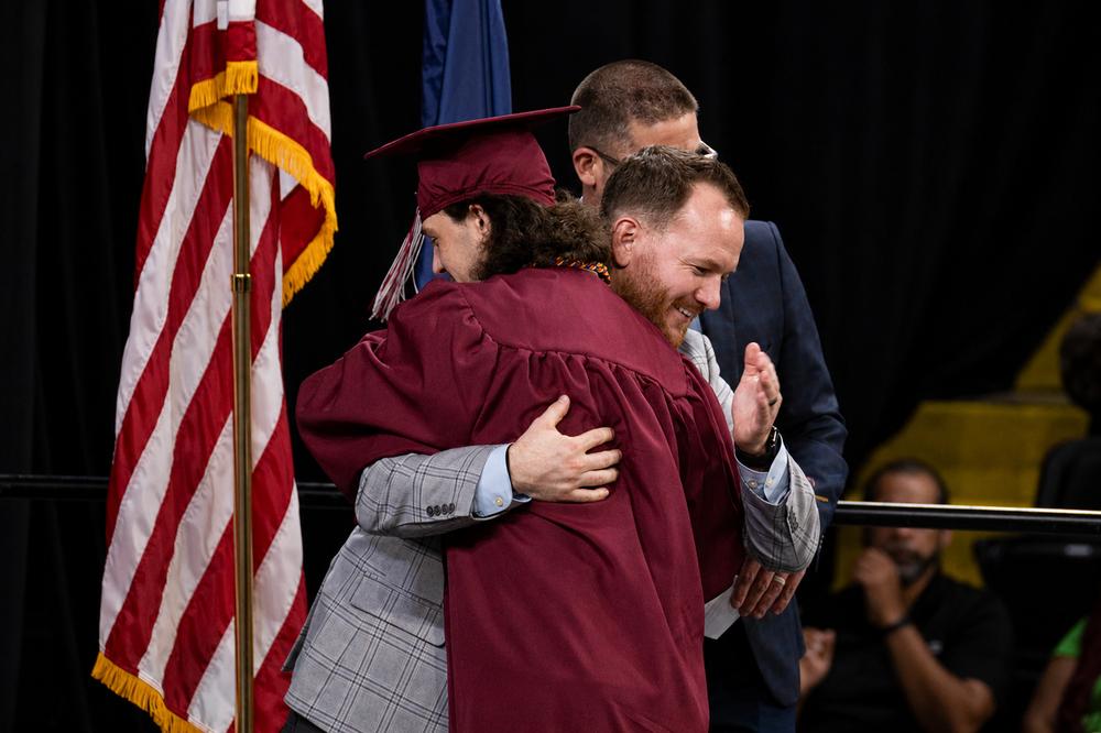Graduate in a maroon cap and gown hugs a district representative on stage during BCTC’s Senior Recognition Ceremony. An American flag stands nearby as others look on.